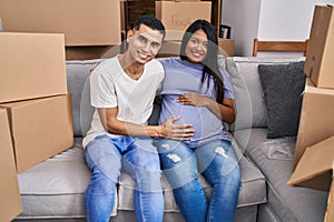 Young hispanic couple expecting a baby sitting on the sofa at new home looking positive and happy standing and smiling with a