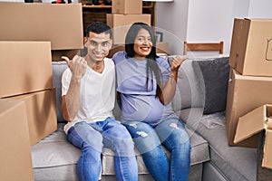 Young hispanic couple expecting a baby sitting on the sofa at new home pointing thumb up to the side smiling happy with open mouth