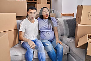 Young hispanic couple expecting a baby sitting on the sofa at new home scared and amazed with open mouth for surprise, disbelief