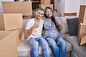 Young hispanic couple expecting a baby sitting on the sofa at new home smiling happy and positive, thumb up doing excellent and