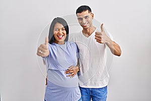 Young hispanic couple expecting a baby standing over background approving doing positive gesture with hand, thumbs up smiling and