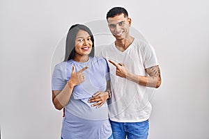 Young hispanic couple expecting a baby standing over background cheerful with a smile on face pointing with hand and finger up to