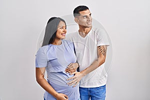 Young hispanic couple expecting a baby standing over background looking away to side with smile on face, natural expression