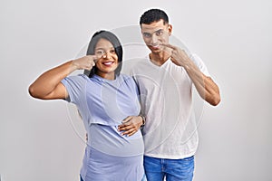 Young hispanic couple expecting a baby standing over background pointing with hand finger to face and nose, smiling cheerful