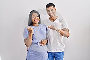 Young hispanic couple expecting a baby standing over background pointing to the back behind with hand and thumbs up, smiling