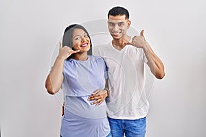 Young hispanic couple expecting a baby standing over background smiling doing phone gesture with hand and fingers like talking on