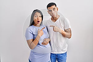 Young hispanic couple expecting a baby standing over background surprised pointing with finger to the side, open mouth amazed