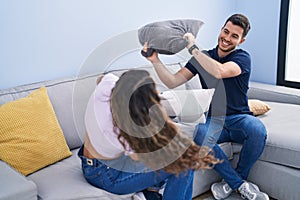 Young hispanic couple fighting with cushion sitting on sofa at home