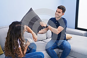 Young hispanic couple fighting with cushion sitting on sofa at home