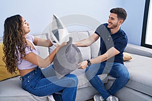 Young hispanic couple fighting with cushion sitting on sofa at home