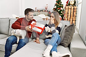 Young hispanic couple holding gift sitting on the sofa with dog at home