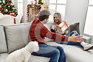 Young hispanic couple holding gift sitting on the sofa with dog at home