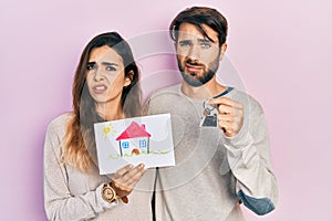 Young hispanic couple holding house draw and key of new home clueless and confused expression
