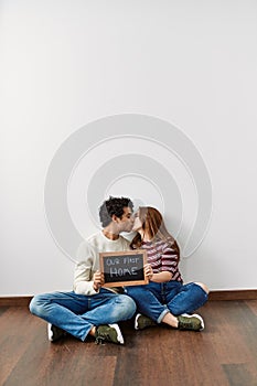 Young hispanic couple holding our first home blackboard sitting on the floor at empty new house