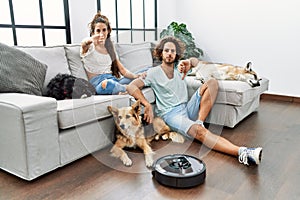 Young hispanic couple relaxing at home by vacuum robot with angry face, negative sign showing dislike with thumbs down, rejection