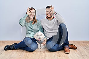 Young hispanic couple sitting on the floor with dog doing ok gesture with hand smiling, eye looking through fingers with happy