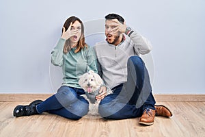 Young hispanic couple sitting on the floor with dog peeking in shock covering face and eyes with hand, looking through fingers