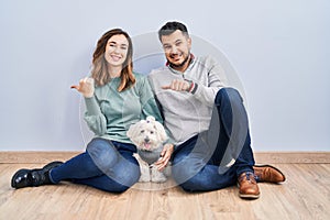 Young hispanic couple sitting on the floor with dog pointing to the back behind with hand and thumbs up, smiling confident