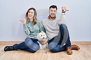 Young hispanic couple sitting on the floor with dog smiling with happy face looking and pointing to the side with thumb up