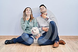 Young hispanic couple sitting on the floor with dog touching painful neck, sore throat for flu, clod and infection