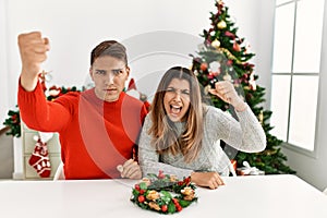 Young hispanic couple sitting at the table on christmas angry and mad raising fist frustrated and furious while shouting with