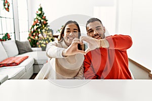 Young hispanic couple sitting on the table by christmas tree smiling in love showing heart symbol and shape with hands