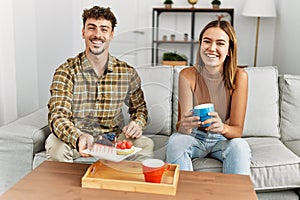 Young hispanic couple smiling happy having breakfast sitting on the sofa at home