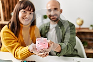 Young hispanic couple smiling happy holding piggy bank sitting on the table at home