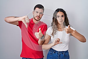 Young hispanic couple standing over isolated background doing thumbs up and down, disagreement and agreement expression