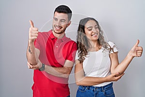 Young hispanic couple standing over isolated background looking proud, smiling doing thumbs up gesture to the side