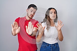 Young hispanic couple standing over isolated background moving away hands palms showing refusal and denial with afraid and
