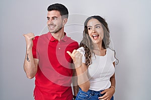 Young hispanic couple standing over isolated background smiling with happy face looking and pointing to the side with thumb up