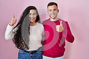 Young hispanic couple standing over pink background doing happy thumbs up gesture with hand