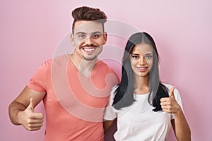 Young hispanic couple standing over pink background doing happy thumbs up gesture with hand