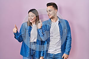 Young hispanic couple standing over pink background looking proud, smiling doing thumbs up gesture to the side