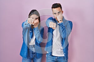 Young hispanic couple standing over pink background punching fist to fight, aggressive and angry attack, threat and violence