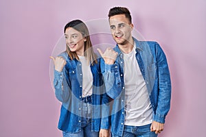 Young hispanic couple standing over pink background smiling with happy face looking and pointing to the side with thumb up