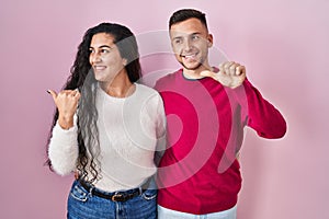 Young hispanic couple standing over pink background smiling with happy face looking and pointing to the side with thumb up