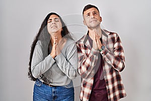 Young hispanic couple standing over white background begging and praying with hands together with hope expression on face very