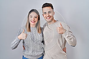 Young hispanic couple standing over white background doing happy thumbs up gesture with hand