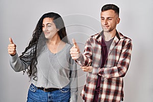 Young hispanic couple standing over white background looking proud, smiling doing thumbs up gesture to the side