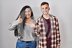 Young hispanic couple standing over white background showing palm hand and doing ok gesture with thumbs up, smiling happy and