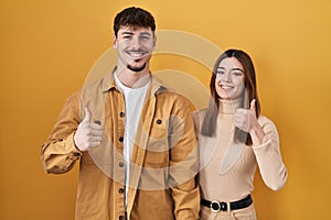 Young hispanic couple standing over yellow background doing happy thumbs up gesture with hand