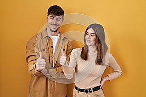 Young hispanic couple standing over yellow background looking proud, smiling doing thumbs up gesture to the side