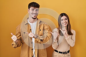 Young hispanic couple standing over yellow background pointing to the back behind with hand and thumbs up, smiling confident