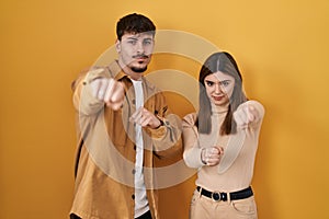 Young hispanic couple standing over yellow background punching fist to fight, aggressive and angry attack, threat and violence