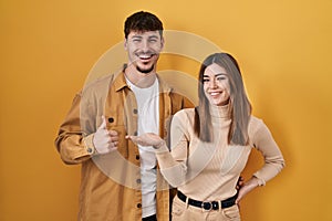 Young hispanic couple standing over yellow background showing palm hand and doing ok gesture with thumbs up, smiling happy and