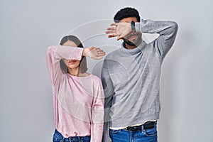 Young hispanic couple standing together covering eyes with arm, looking serious and sad