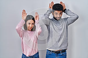 Young hispanic couple standing together doing bunny ears gesture with hands palms looking cynical and skeptical