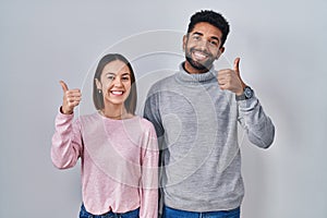 Young hispanic couple standing together doing happy thumbs up gesture with hand